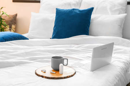 Tray with cup and earphones, laptop on soft bed