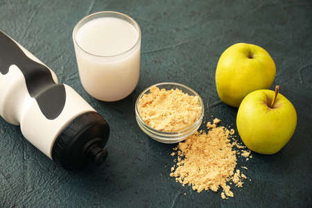 Glass of protein shake, bowl with powder, bottle and apples on dark table