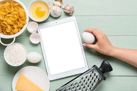 Woman holding egg, digital recipe book and ingredients on color wooden backgroundの写真素材
