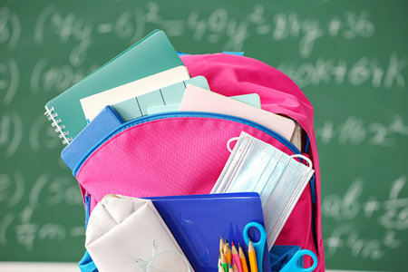 School backpack with stationery and medical mask against blackboard, closeupの写真素材