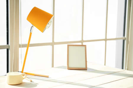 Modern lamp, cup and blank photo frame on white wooden table near big windowの写真素材