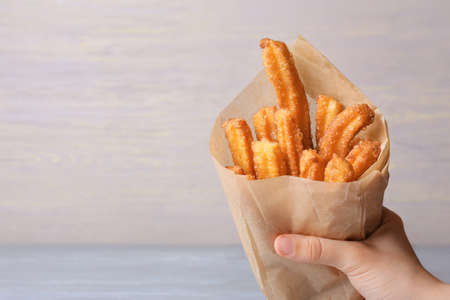 Female hand with tasty churros in parchment on light wooden background, closeup