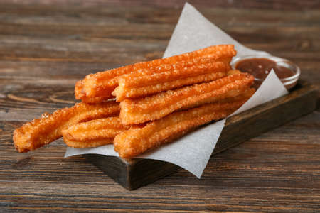 Board with tasty churros and melted chocolate sauce in bowl on wooden background, closeupの写真素材