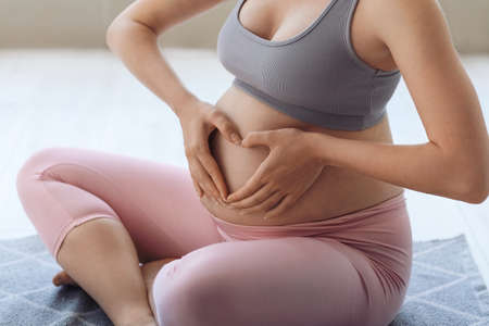Young pregnant woman making heart with her hands at home, closeup