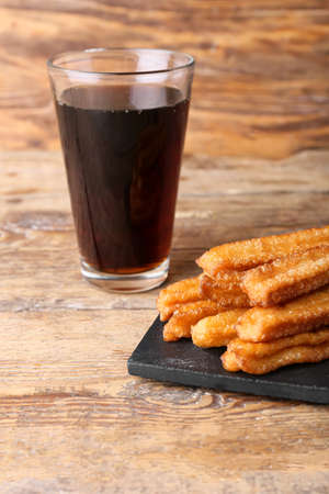 Board with tasty churros and glass of cola on wooden background
