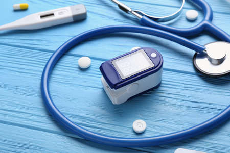 Pulse oximeter with pills and stethoscope on color wooden background, closeup