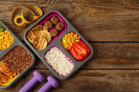 Containers with healthy food, dumbbells and measuring tape on wooden background, closeup