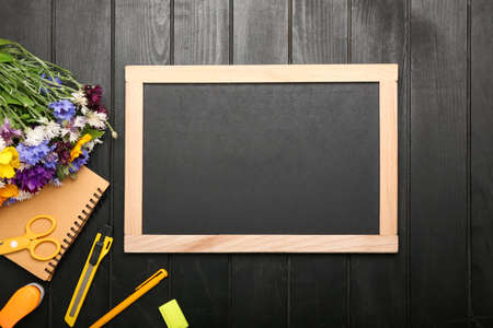 Blank chalkboard, stationery and flowers on dark wooden background, closeup