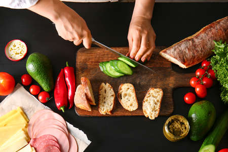 Woman cutting cucumber for tasty sandwiches on dark background