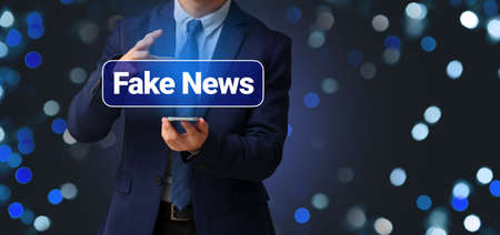 Businessman with smartphone and text FAKE NEWS on virtual screen against dark backgroundの写真素材