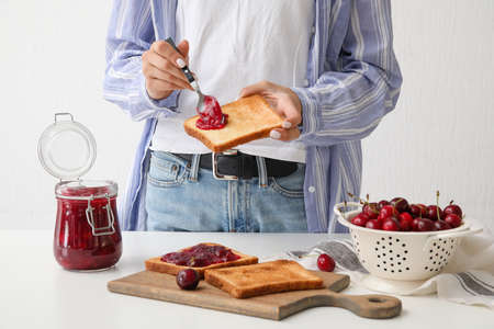 Woman spreading sweet cherry jam onto toast in kitchen