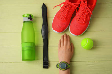 Female hand with fitness tracker, chest belt, bottle of water, tennis ball and sport shoes on color...