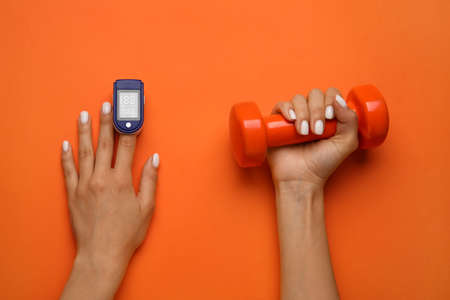 Female hands with pulse oximeter and dumbbell on color backgroundの写真素材