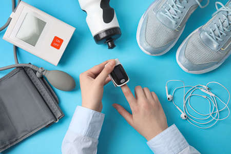 Female hands with pulse oximeter, sphygmomanometer, earphones, bottle of water and sport shoes on color backgroundの写真素材