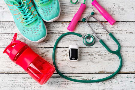 Pulse oximeter, stethoscope, bottle of water, shoes and skipping rope on light wooden background