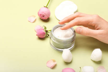 Female hand with jar of cosmetic product and rose flowers on green background, closeup