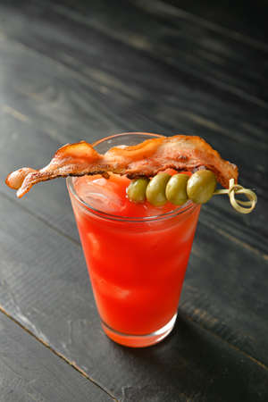 Glass of bloody mary cocktail garnished with bacon and olives on dark wooden backgroundの写真素材