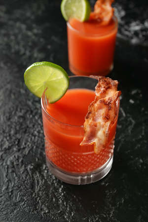 Glasses of bloody mary cocktail garnished with bacon and lime on dark background