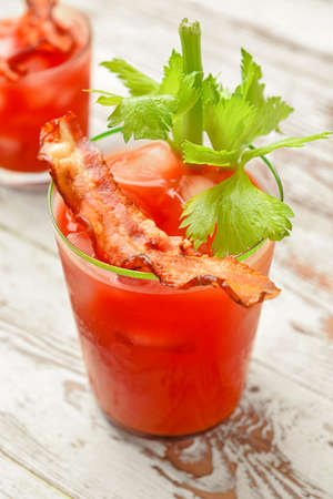 Glass of bloody mary cocktail garnished with bacon and cilantro on light wooden background