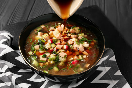 Pouring of kvass into bowl with okroshka on dark wooden background, closeup