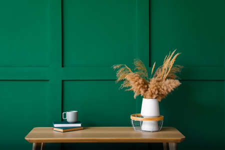 Vase with dry reeds, cup and books on bench near green wall