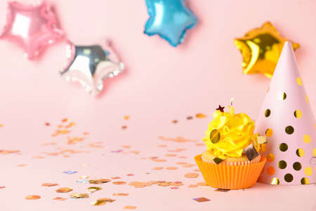 Tasty Birthday cupcake with burning candle and hat on color background