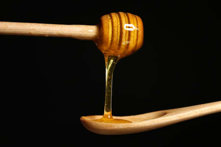 Honey dripping from dipper into wooden spoon on black background, closeup