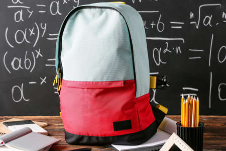 School backpack with stationery on table against blackboard