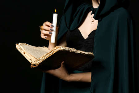 Young witch with spell book and candle on dark background