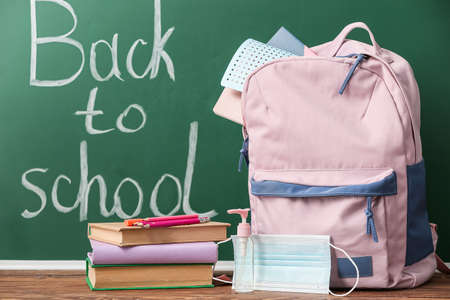 School backpack with stationery, medical mask and bottle of sanitizer on table in classroom