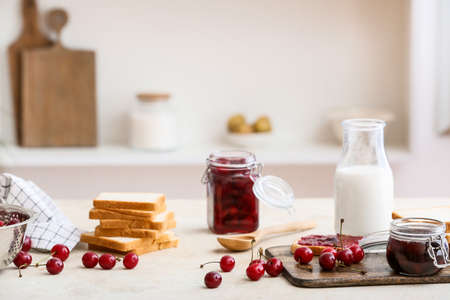 Jar with tasty cherry jam, bread and bottle of milk on table in kitchenの写真素材