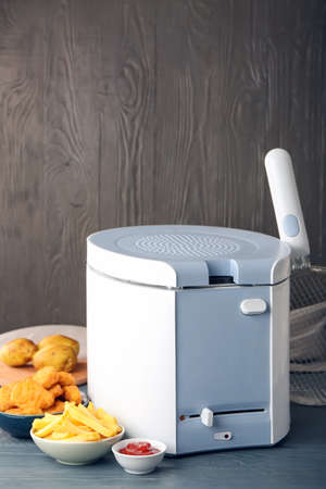 Deep fryer with bowls of delicious fried potatoes and nuggets on wooden background