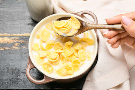 Woman preparing tasty cornflakes with milk on dark wooden background, closeup