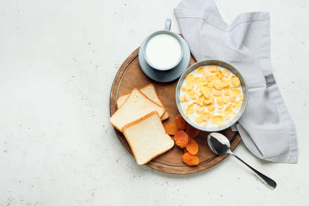Bowl with tasty cornflakes, cup of milk, bread and dried apricots on light background