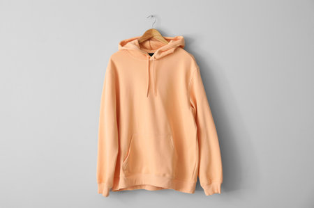 Stylish peachy hoodie hanging on light wall