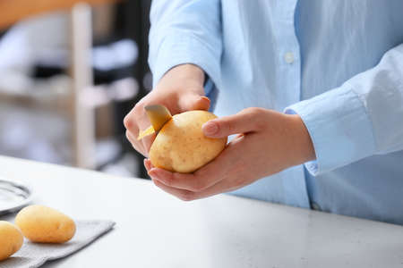 Woman peeling raw potatoes at table in kitchen, closeup