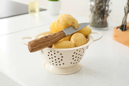 Colander with raw potatoes and knife on table at kitchenの写真素材