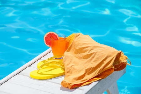 Swimsuit, towel, flip-flops and tasty cocktail on wooden table near swimming pool