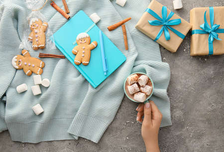 Female hand with cup of cacao, cookies, gifts, notebook and sweater on grunge background