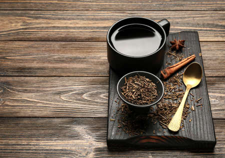 Board with dry hojicha green tea and cup of hot beverage on wooden background