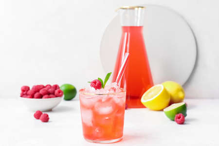 Glass of tasty lemonade and bowl with raspberry on light background