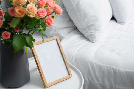 Vase with bouquet of beautiful fresh roses and blank frame on table in bedroom, closeup