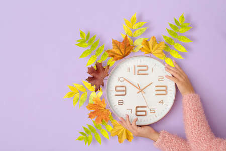 Female hands with clock and autumn leaves on lilac background. daylight saving time