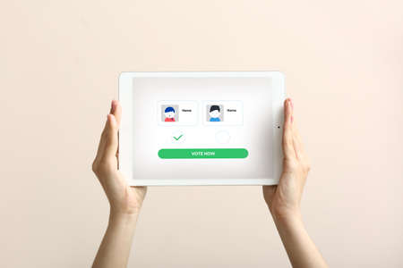 Female hands holding tablet computer with text VOTE NOW and icons of candidates on screen against light backgroundの写真素材