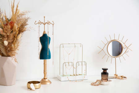 Stands with stylish jewelry, vase with flowers, mirror and perfume bottle on chest of drawers near...