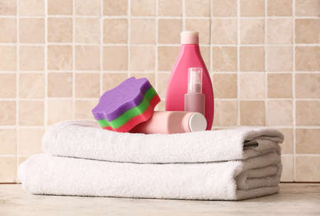 Stack of clean towels, bottles of cosmetic products and bath sponge on table near color tileの写真素材