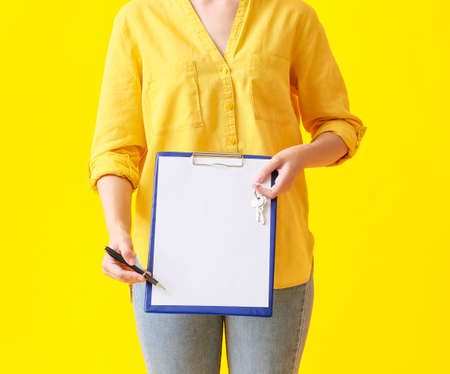 Young woman with keys from house and clipboard on yellow backgroundの写真素材