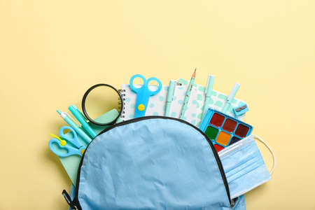 Backpack with set of stationery and medical mask on color background