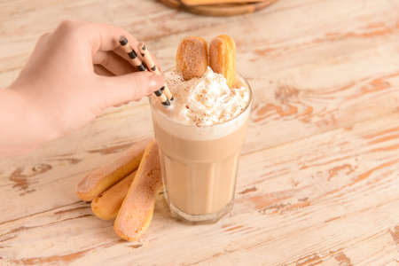 Female hand and glass of tasty Tiramisu Latte on white wooden background