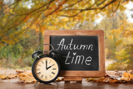 Alarm clock, chalkboard with text AUTUMN TIME and dry leaves on table outdoorsの写真素材